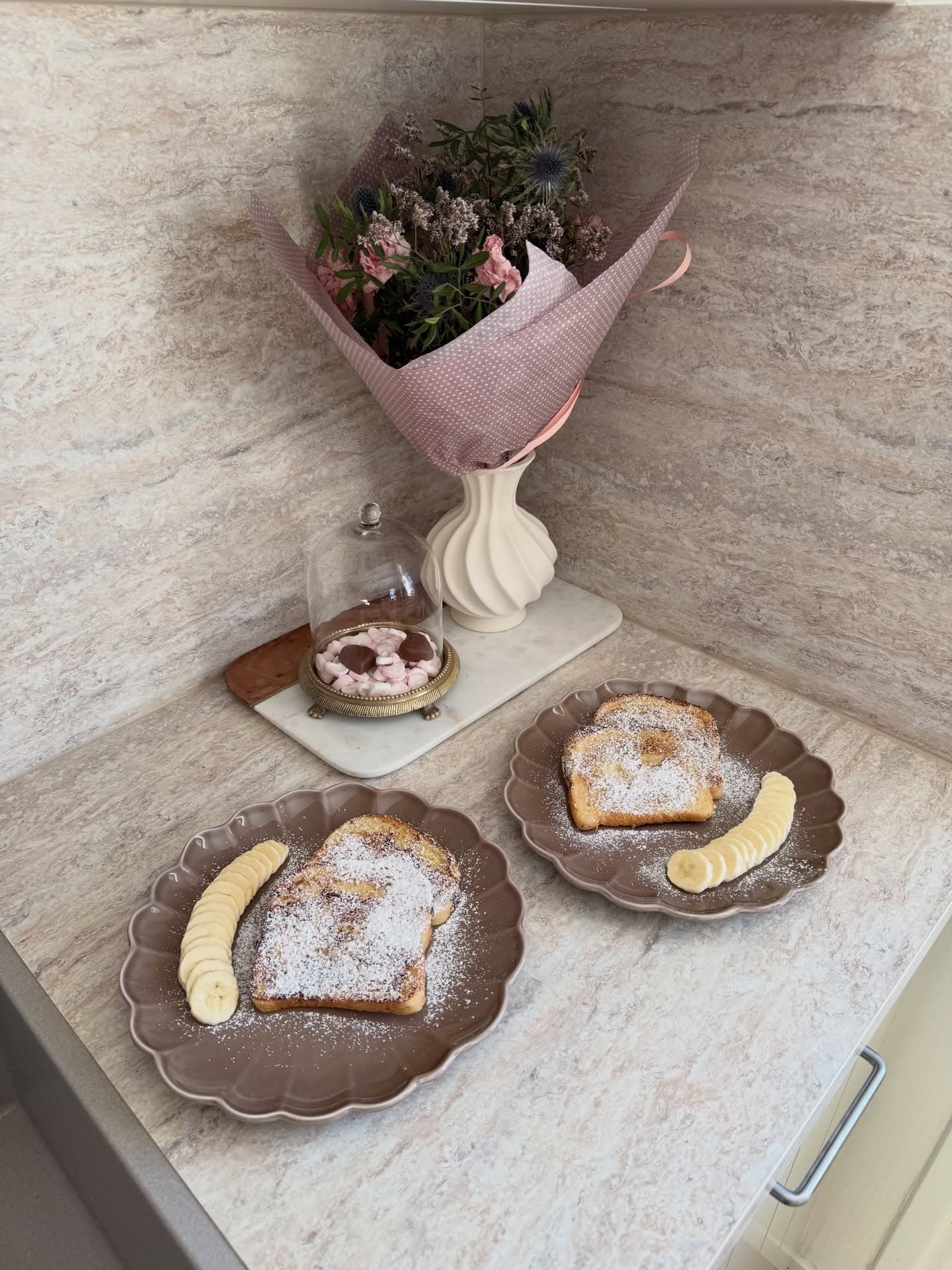 Two plates of French toast topped with powdered sugar and banana slices, with a bouquet in a vase behind.
